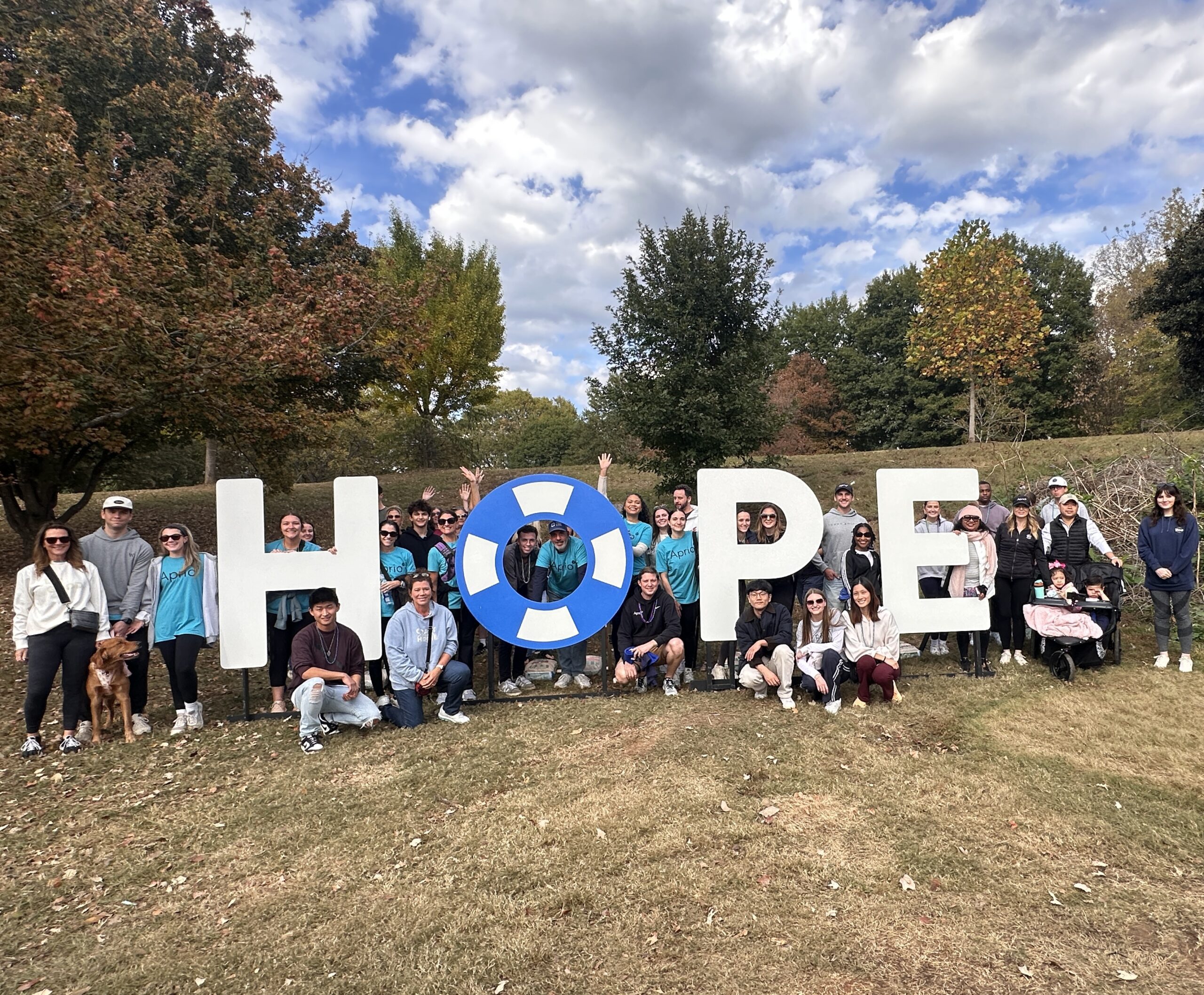 A group of people pose outdoors around large letters spelling HOPE, with trees and a partly cloudy sky in the background.