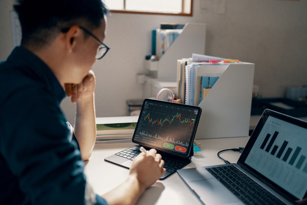A person looks at a tablet displaying a financial chart with a rising trend, while a laptop beside them shows a bar graph.