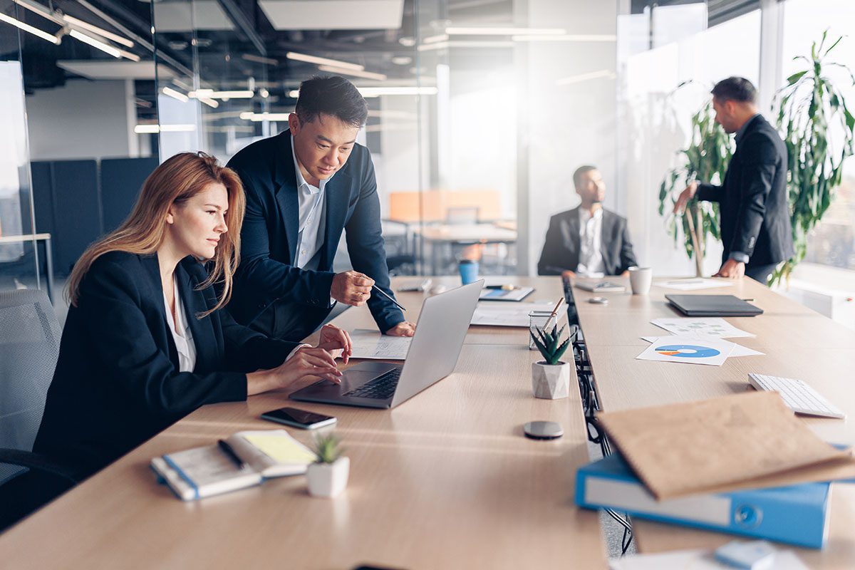 Two colleagues in business attire work on a laptop at a conference table, while two others converse in the background of a modern office.