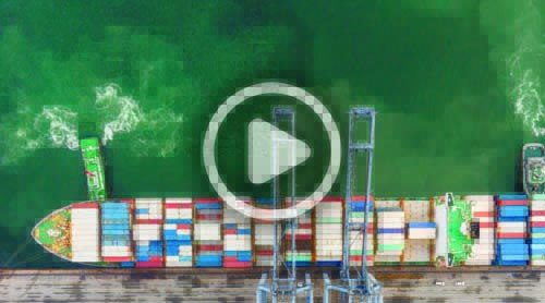 Aerial view of a container ship docked at a port with cranes for loading and unloading; a smaller boat is nearby.