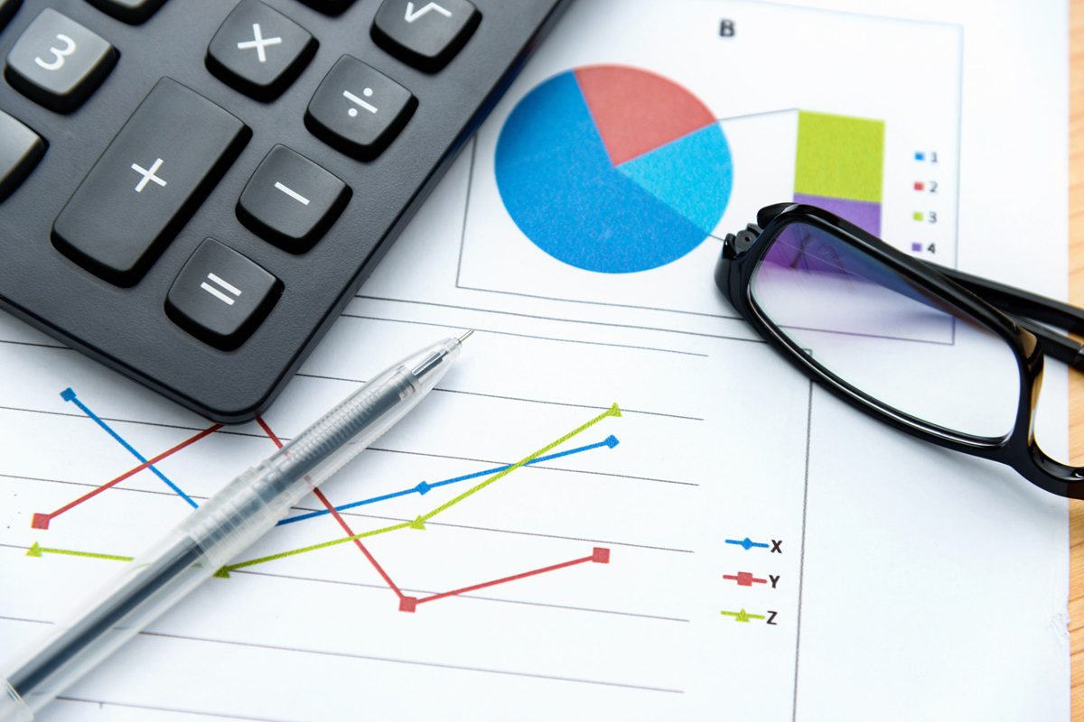 A calculator, eyeglasses, and pen rest on printed charts with colored graphs and pie charts on a desk.