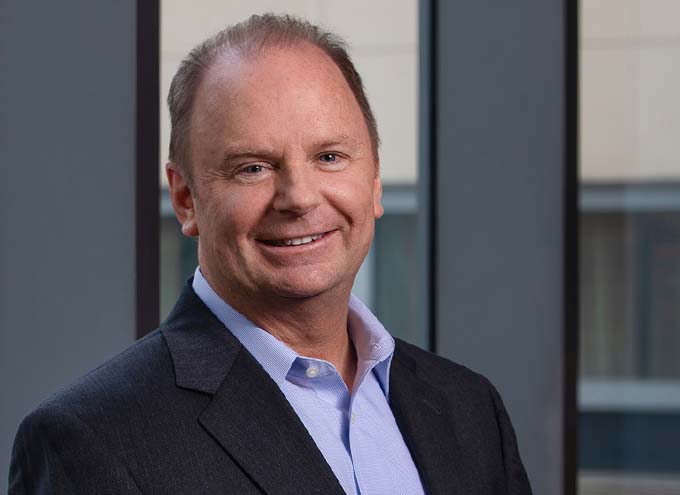 A middle-aged man in a suit jacket and light blue shirt smiles while standing in front of large windows in an office setting.