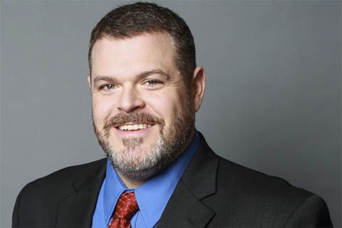 Man with short dark hair and beard wearing a dark suit, blue shirt, and red patterned tie, smiling in front of a plain gray background.