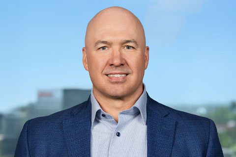 A bald man in a blue suit jacket and light blue shirt stands in front of a blurred outdoor background.