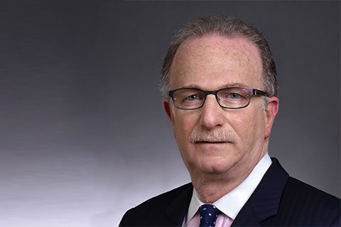 A middle-aged man with glasses, short gray hair, and a mustache, wearing a dark suit, white shirt, and polka dot tie, poses against a gray background.