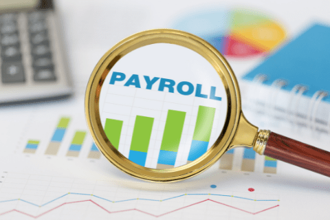 A magnifying glass focuses on the word PAYROLL over a bar graph, with office supplies and financial documents in the background.