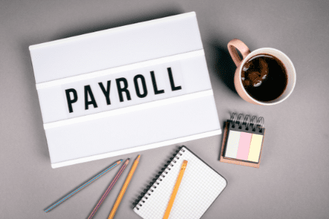 A sign with the word PAYROLL, a cup of coffee, colored pencils, a notepad, and a small calendar on a gray surface.