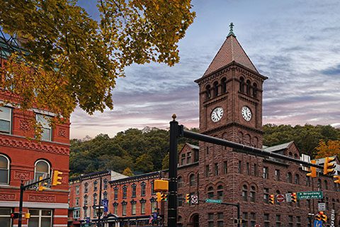 A historic brick clock tower stands at a street intersection lined with brick buildings and traffic lights, with trees and hills in the background.