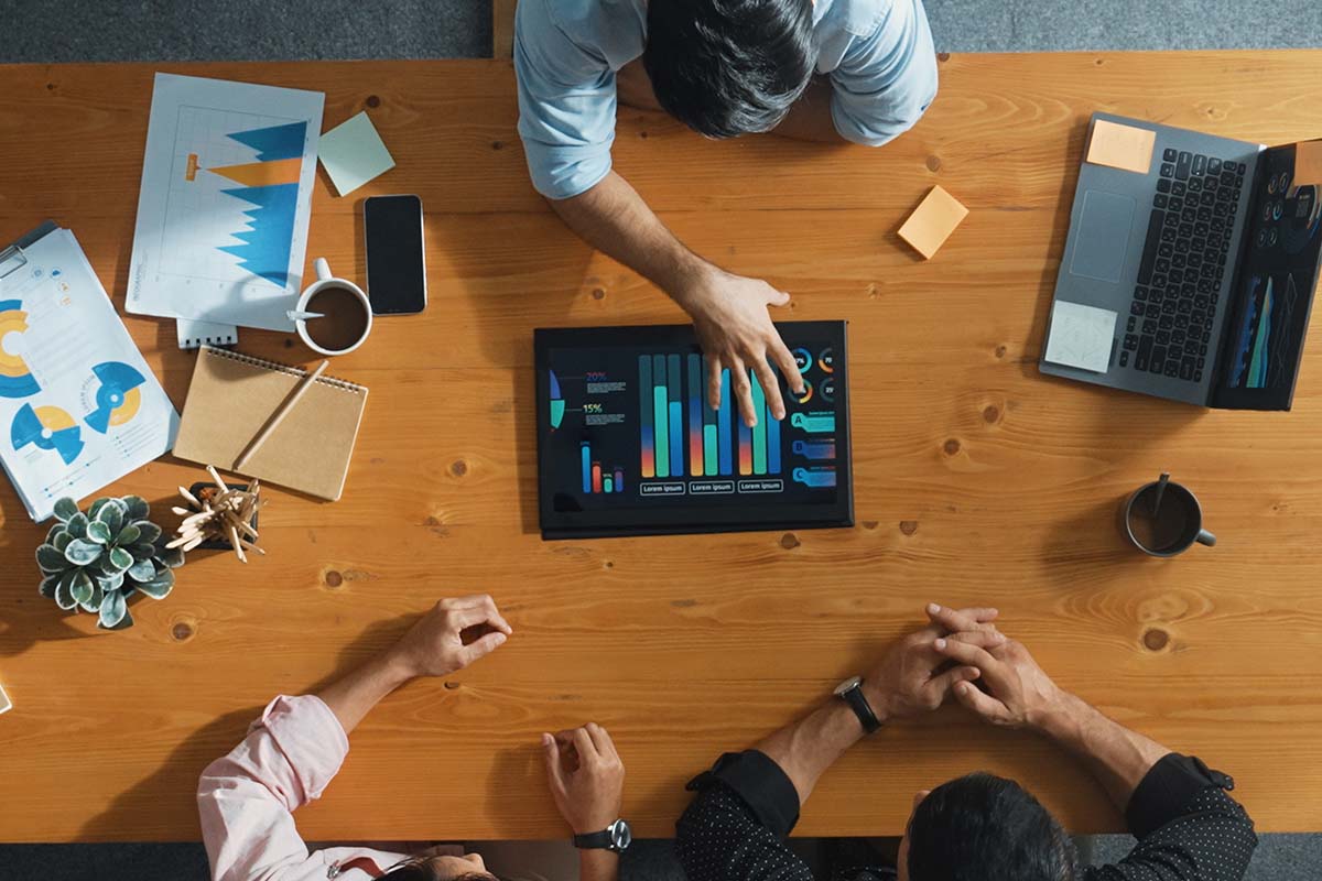 Three people sit around a wooden table reviewing colorful data charts on a tablet, with papers, notebooks, a laptop, and coffee cups also on the table.