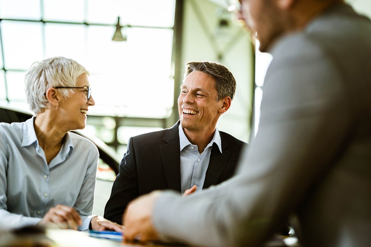 Three business professionals in formal attire sit together at a table, smiling and engaged in conversation in a bright office setting.