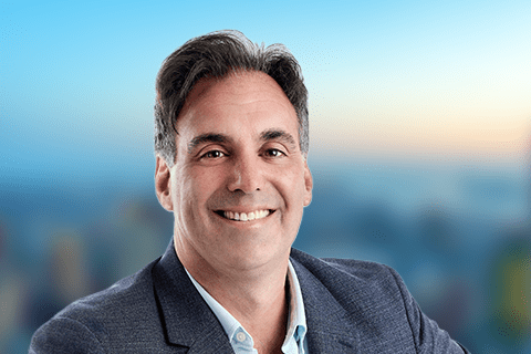 Smiling man in a blue blazer and light blue shirt against a blurred background with a gradient sky.
