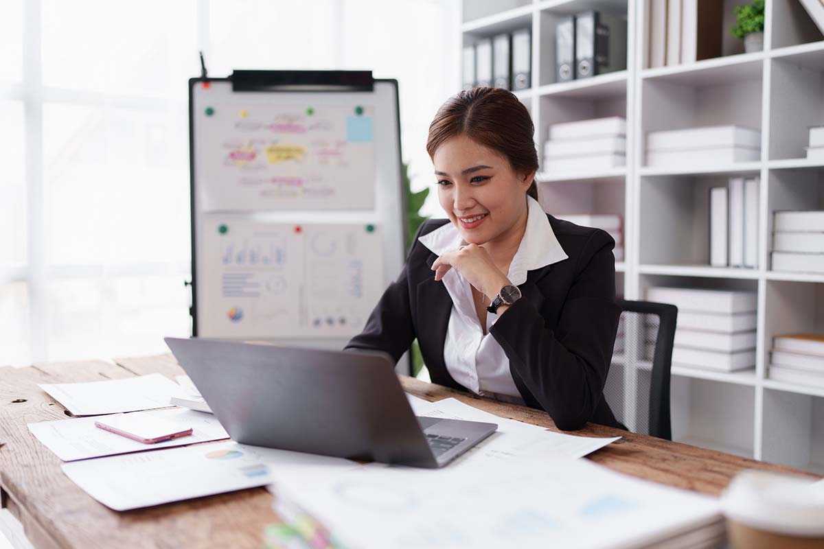 A woman in business attire sits at a desk with papers and a laptop, working and smiling, with charts and graphs displayed on a whiteboard behind her.