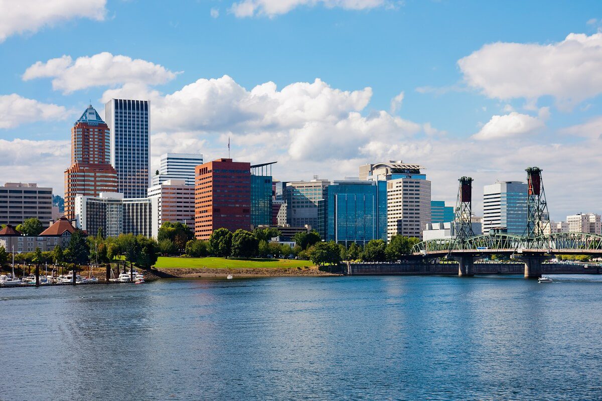 A city skyline with various modern buildings, a waterfront, greenery, and a bridge spanning the river under a partly cloudy sky.