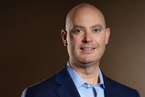 A bald man in a blue suit jacket and light blue shirt poses against a plain brown background, looking at the camera and smiling slightly.