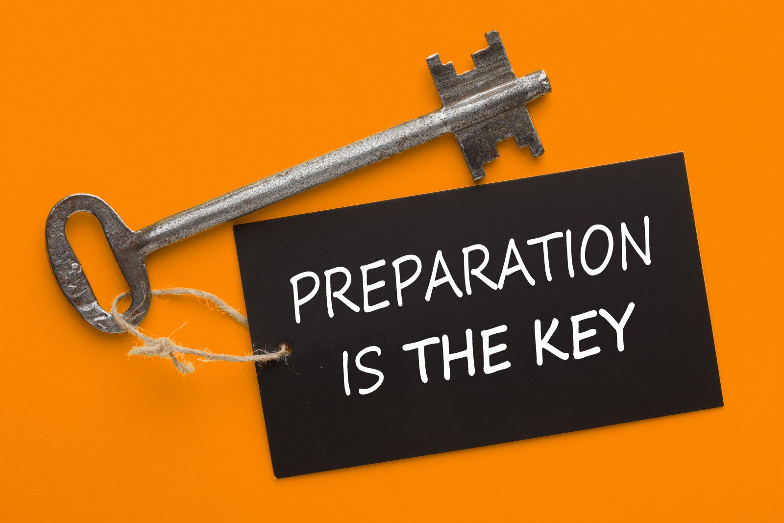 A large metal key lies next to a black card with the words PREPARATION IS THE KEY against an orange background.
