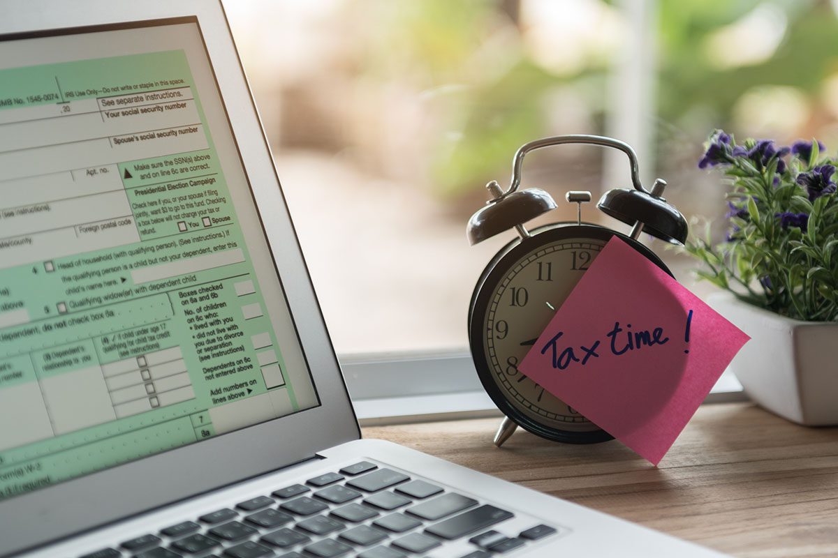 A laptop displaying a tax form sits next to an alarm clock with a pink sticky note reading Tax time! and a small potted plant on a desk.