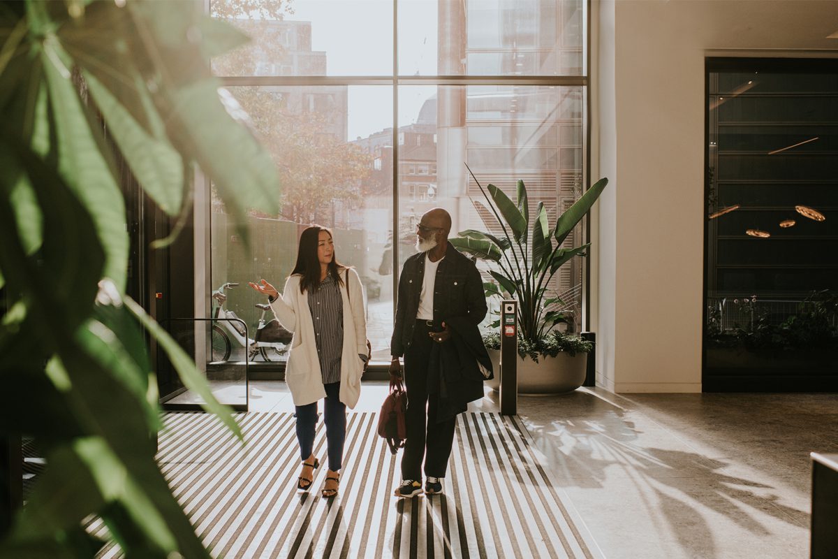 Two people walk and talk in a modern indoor space with large windows, sunlight streaming in, and green plants along the sides.