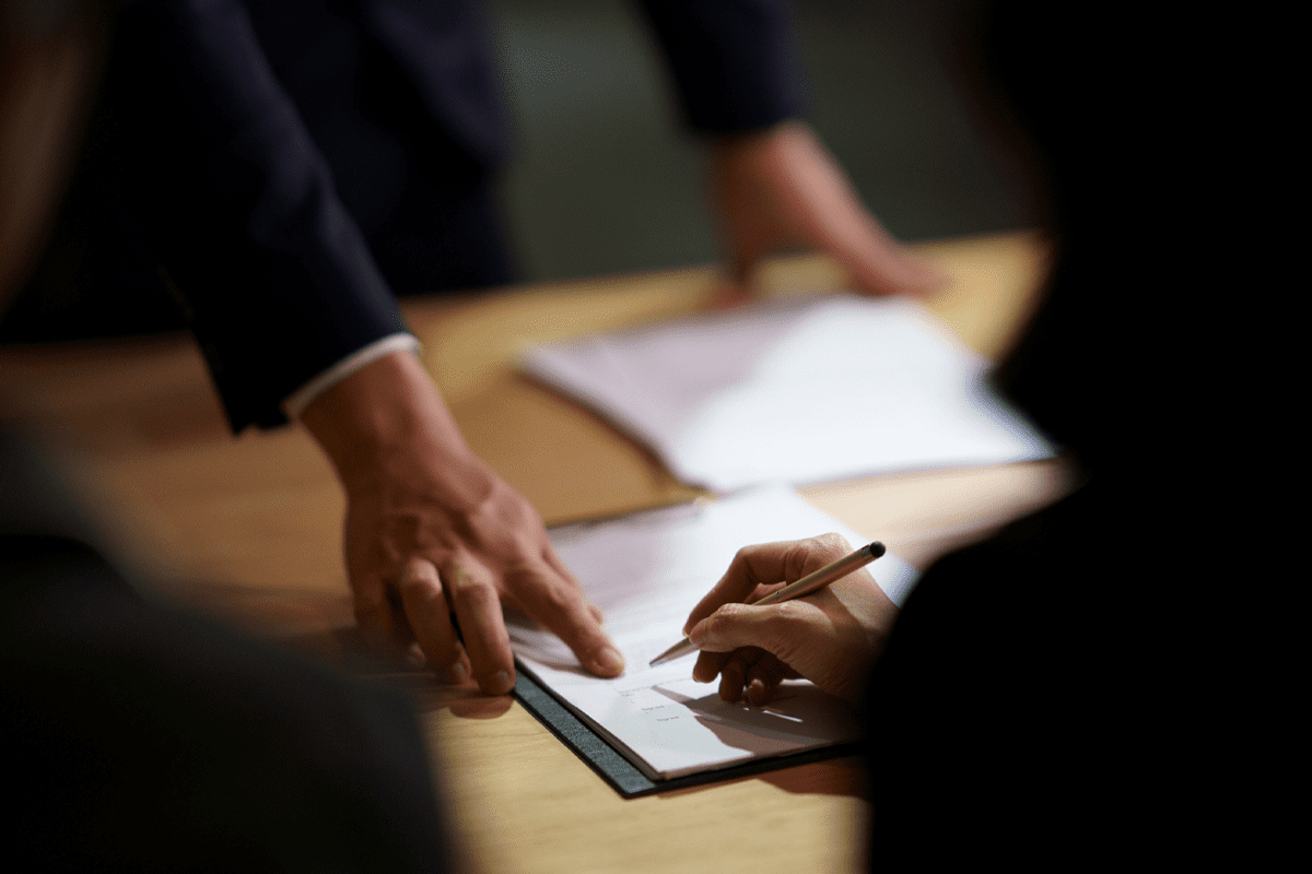 A person signs a document on a clipboard while another person points to the paper on a table with more documents.
