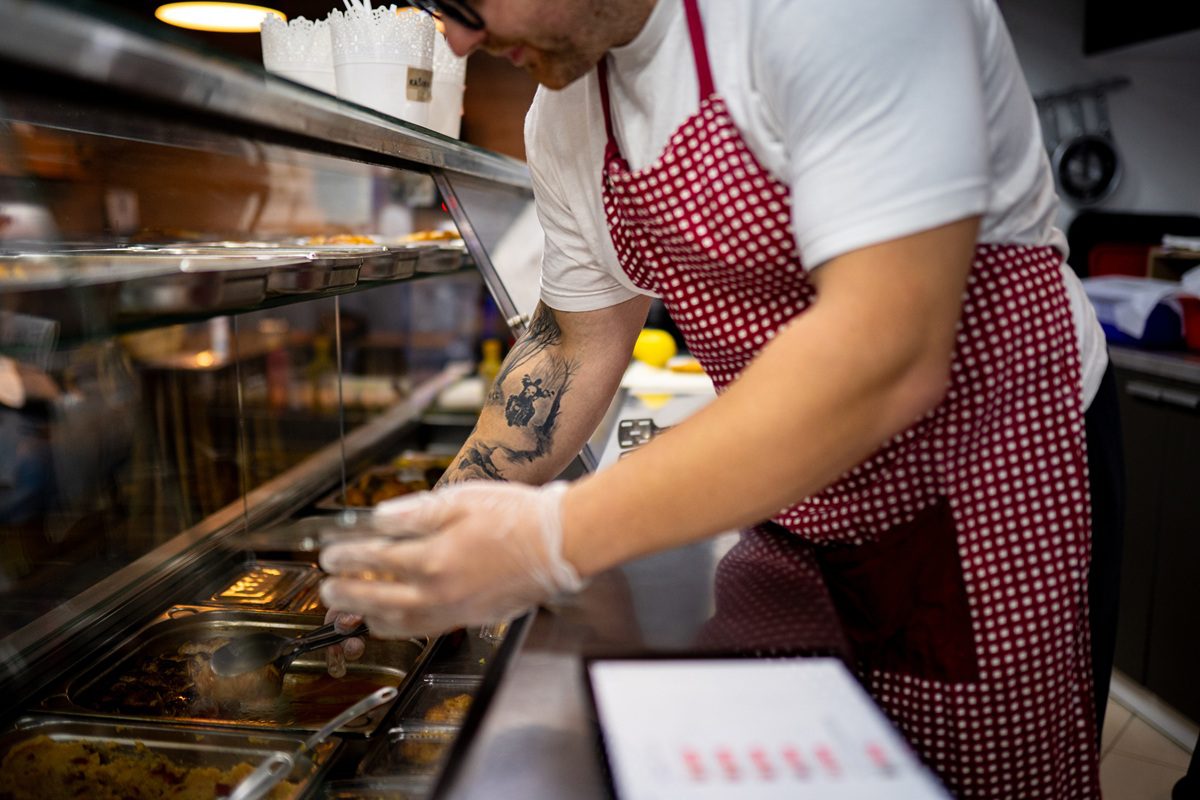 A person wearing a red checkered apron and gloves serves food from a display counter in a restaurant or cafeteria.