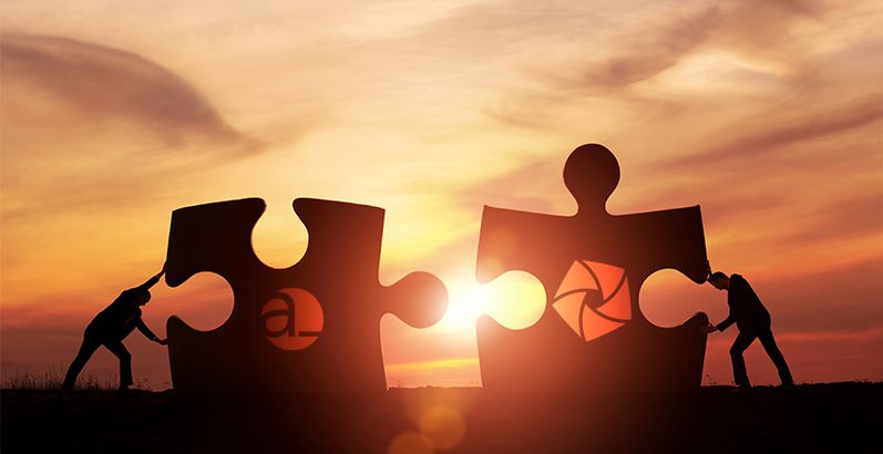 Two silhouetted figures push large puzzle pieces together at sunset, each piece displaying a different logo.