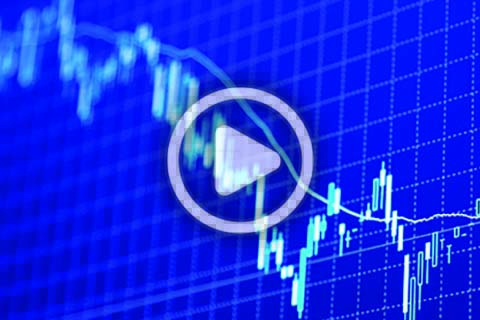 A digital screen displays a fluctuating financial chart with a play button overlay, indicating a video about market data or stock trends.