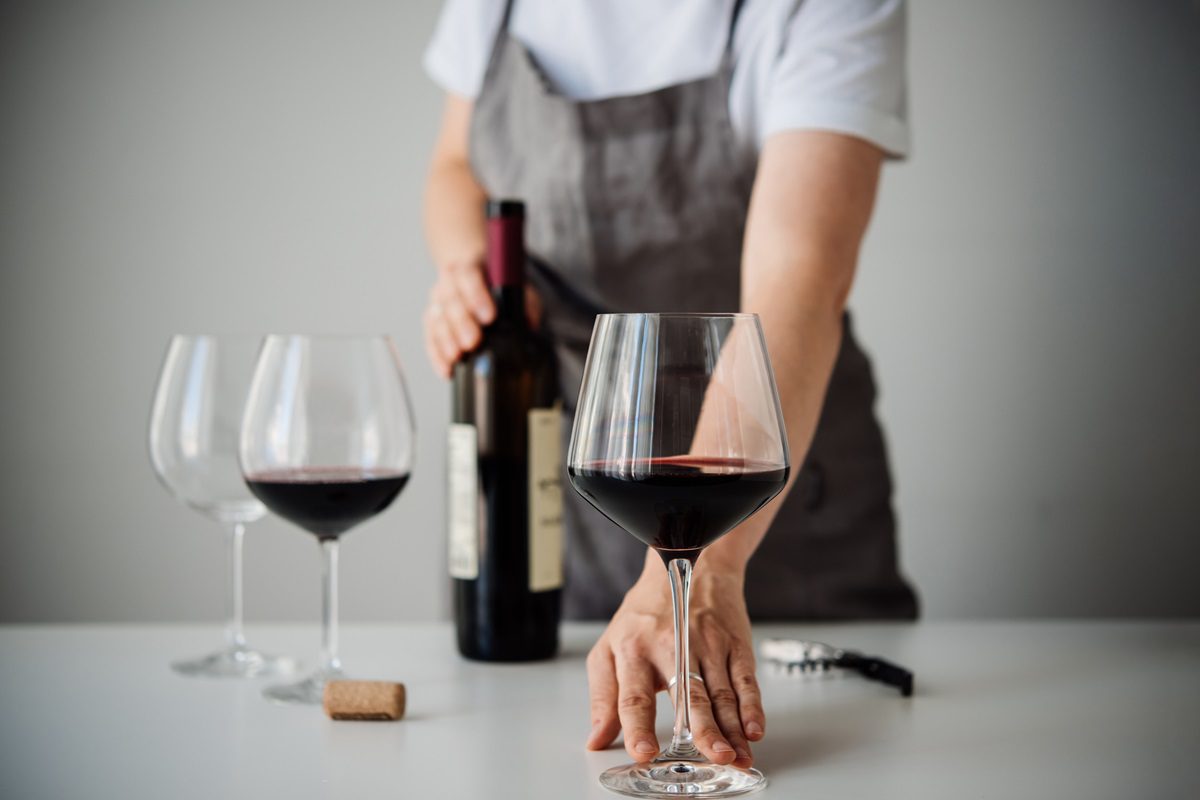 A person in an apron holds a glass of red wine on a table, with two empty glasses, a wine bottle, a cork, and a corkscrew nearby.