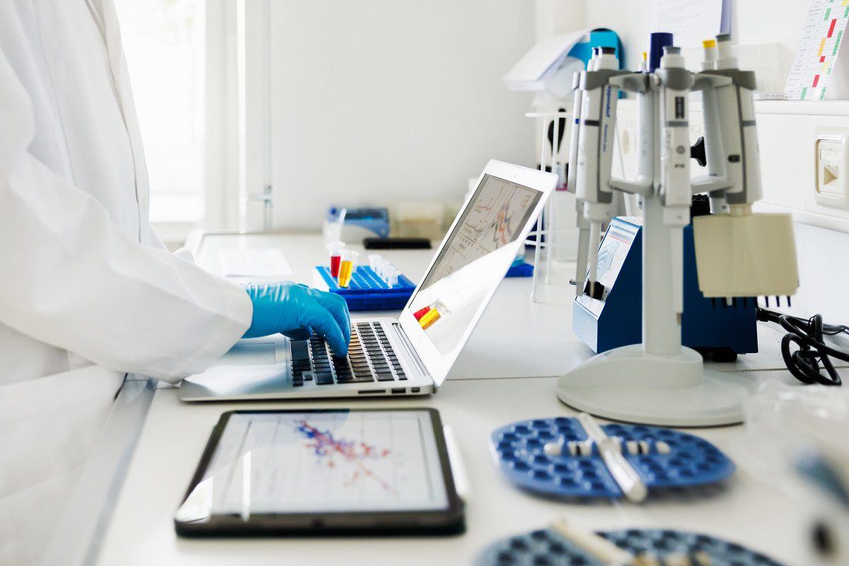 A person in a lab coat and blue gloves works on a laptop in a laboratory, surrounded by scientific equipment, charts, and research materials.