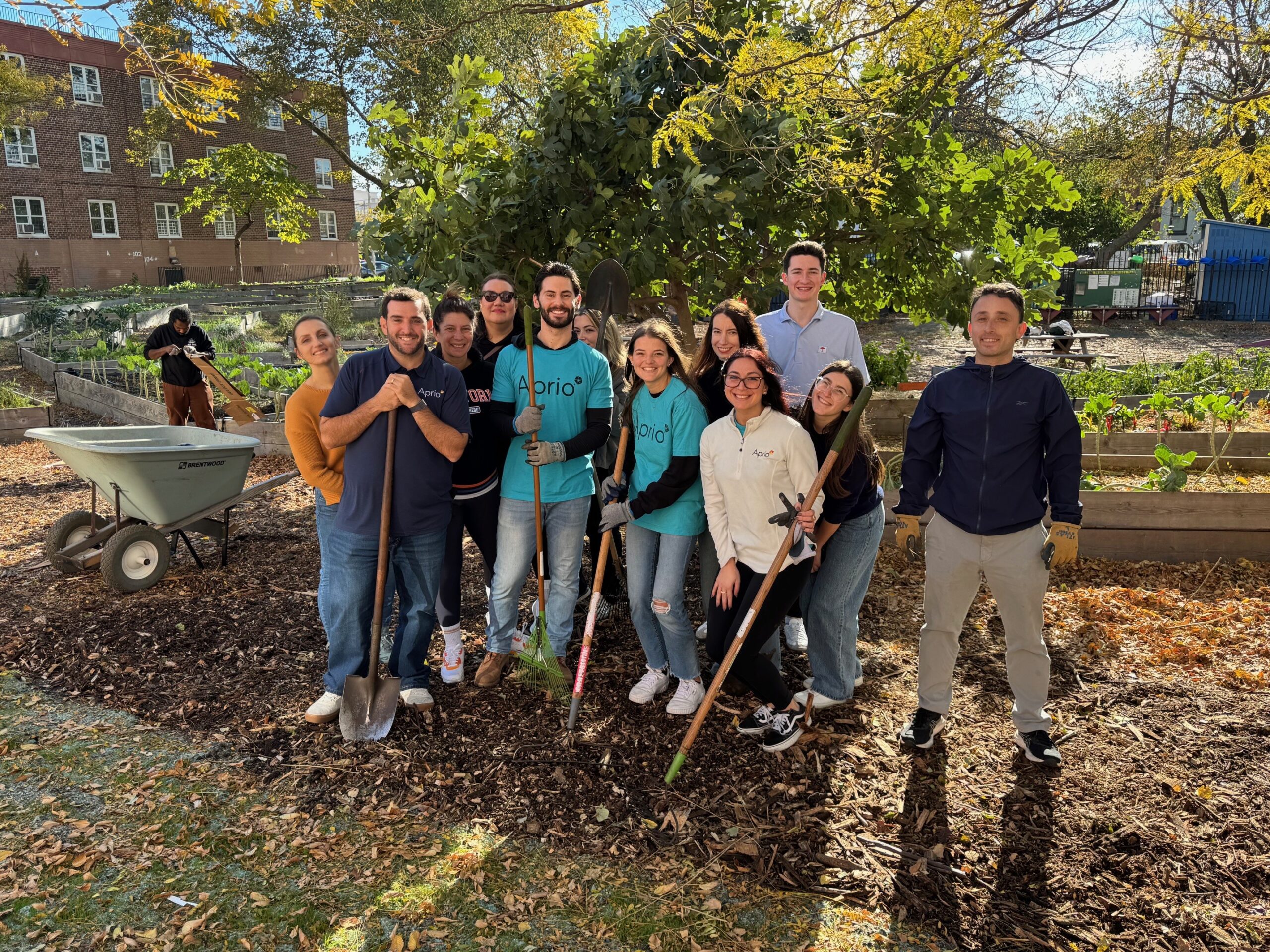 A group of people pose together outdoors in a community garden, holding gardening tools and wearing casual clothing, with trees and garden beds in the background.