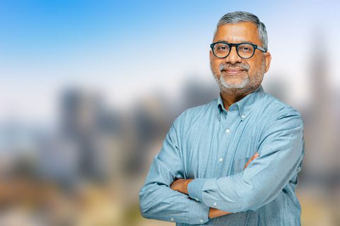 A middle-aged man with gray hair and beard, wearing glasses and a blue shirt, stands with arms crossed in front of a blurred cityscape background.