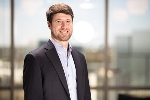 A man in a dark suit and light blue shirt stands smiling in front of large office windows with a blurred cityscape in the background.