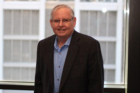 An older man wearing glasses, a blue shirt, and a dark blazer stands indoors in front of a large window with a building visible outside.