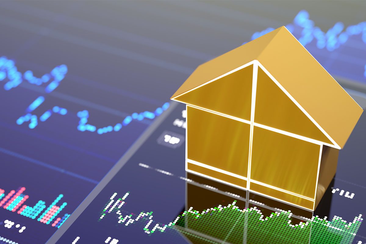 A gold house model sits on a reflective surface displaying financial graphs and stock market data on a blue digital screen.