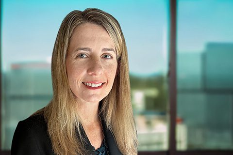A woman with long blonde hair wearing a black blazer stands indoors in front of large windows with an urban background.