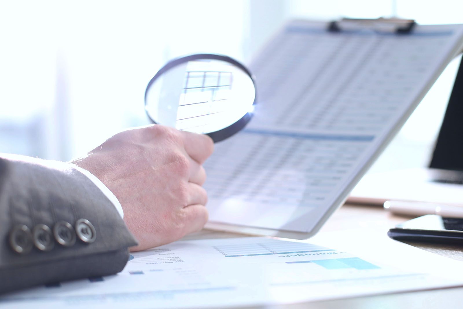 A person holds a magnifying glass over a document on a clipboard, examining printed data in an office setting.