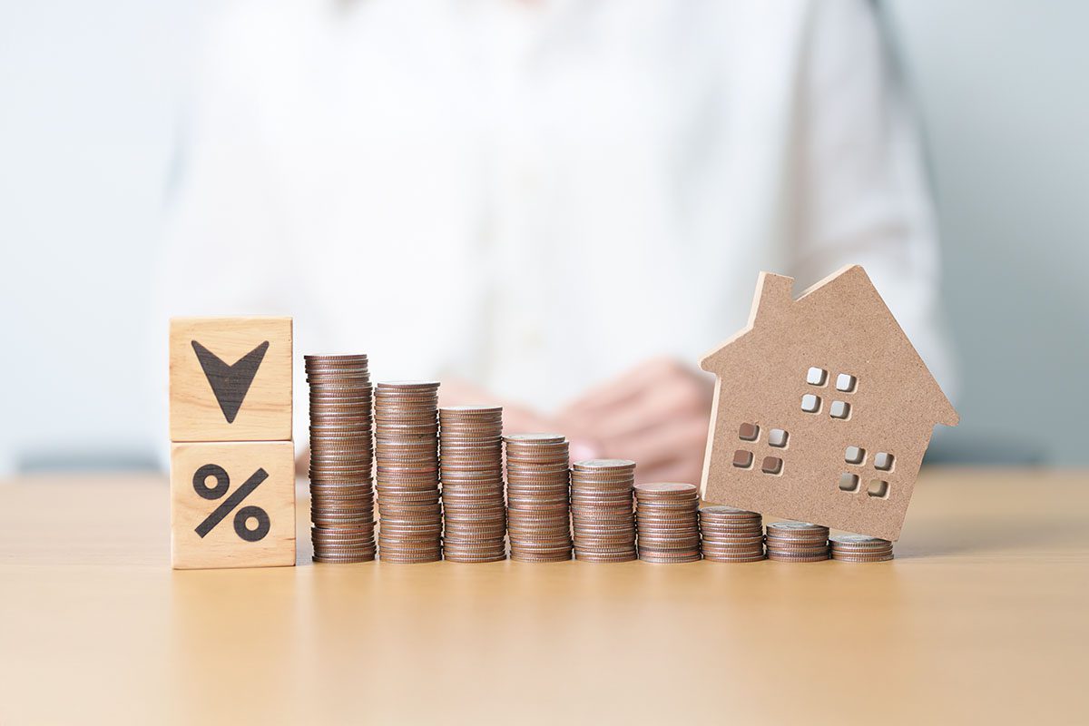 Stacks of coins decrease in height from left to right, with wooden blocks showing a downward arrow and percent sign, and a small house model on the right.
