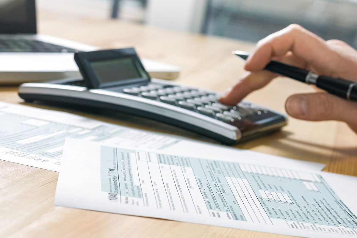 A person uses a calculator while filling out tax forms on a desk with a laptop in the background.
