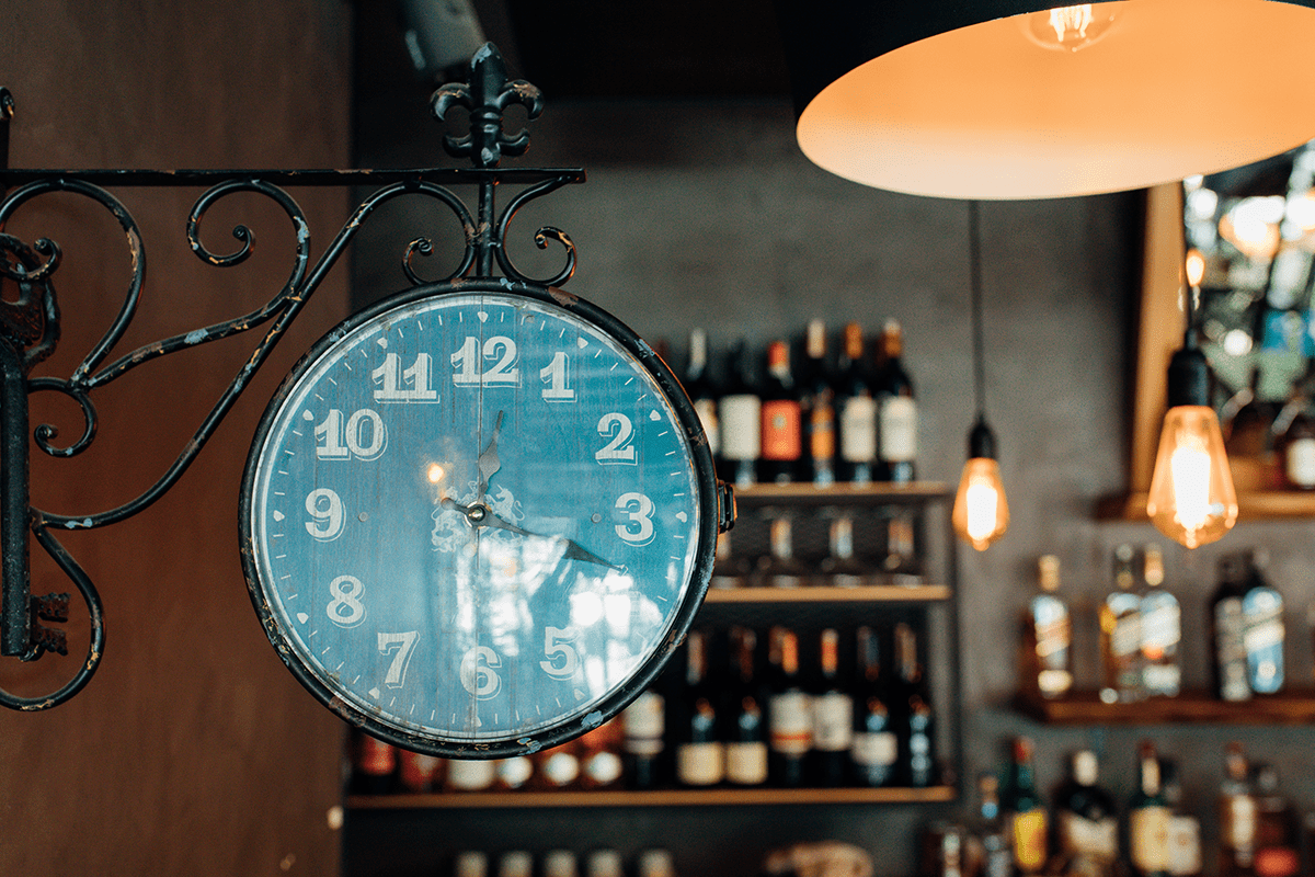 A vintage wall clock shows 9:22 in a bar or restaurant with shelves of bottles and hanging light bulbs in the background.