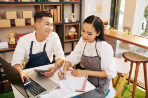 Two people wearing aprons work at a table with a laptop and documents. One is using a calculator, and the other is writing notes. Shelves and a window are in the background.