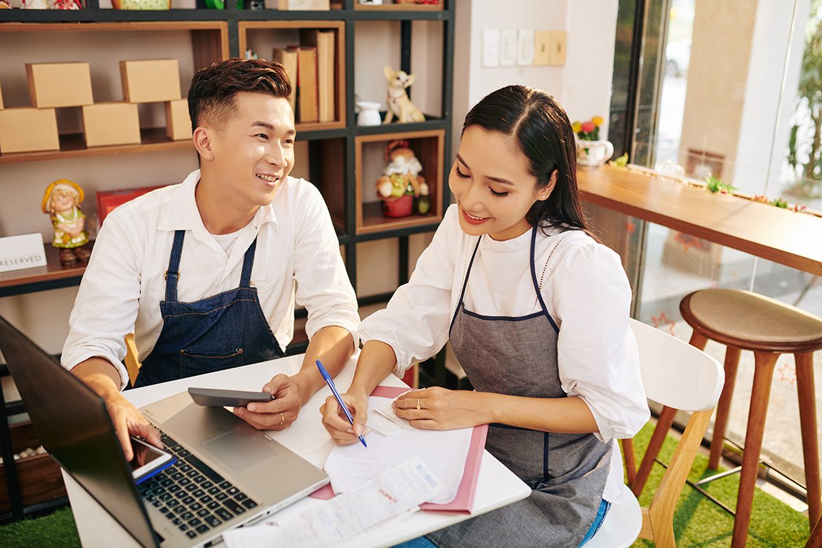 Two people wearing aprons work at a table with a laptop and documents. One is using a calculator, and the other is writing notes. Shelves and a window are in the background.
