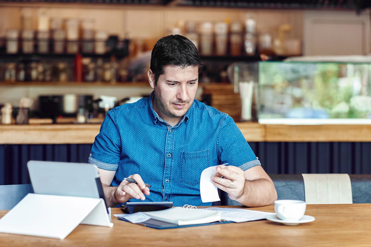 A man sits at a table in a cafe, looking at a receipt and using a calculator, with a tablet, notebook, and a cup of coffee in front of him.