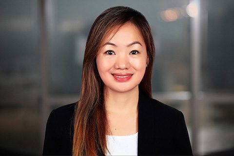 A woman with long straight hair and a black blazer smiles at the camera. The background is blurred, showing large windows.