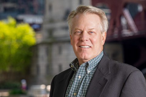 Middle-aged man with light hair wearing a plaid shirt and dark blazer standing outdoors in front of a blurred urban background.