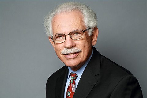 Older man with white hair, glasses, and a mustache wearing a dark suit, light blue shirt, and patterned tie, posed against a plain gray background.