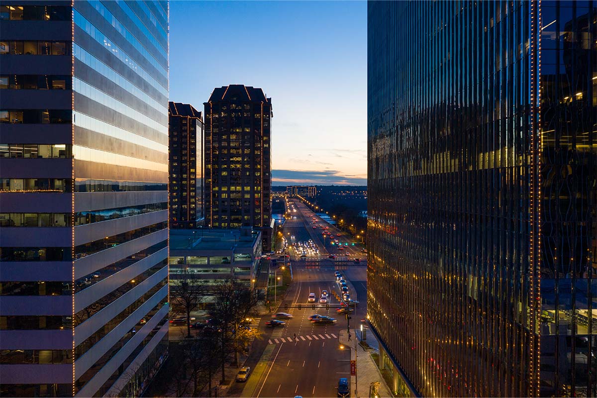 A city street at dusk is lined with tall office buildings, with cars and streetlights visible, and a fading sunset in the background.