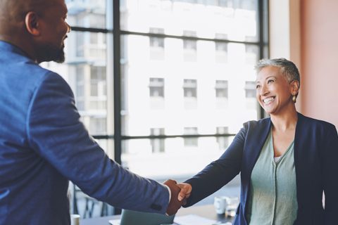 Two people in business attire shake hands and smile at each other in a bright office setting with large windows in the background.