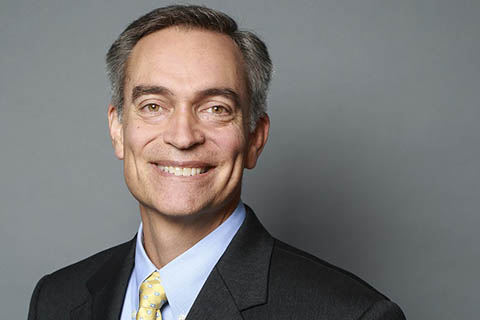 A middle-aged man in a dark suit, light blue shirt, and yellow polka dot tie smiles at the camera against a plain gray background.