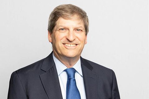 A man in a dark suit and blue tie smiles at the camera against a plain light background.