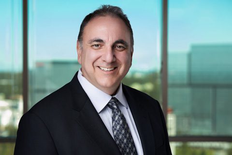 A man in a dark suit and patterned tie smiles while standing in front of large office windows with an urban background.