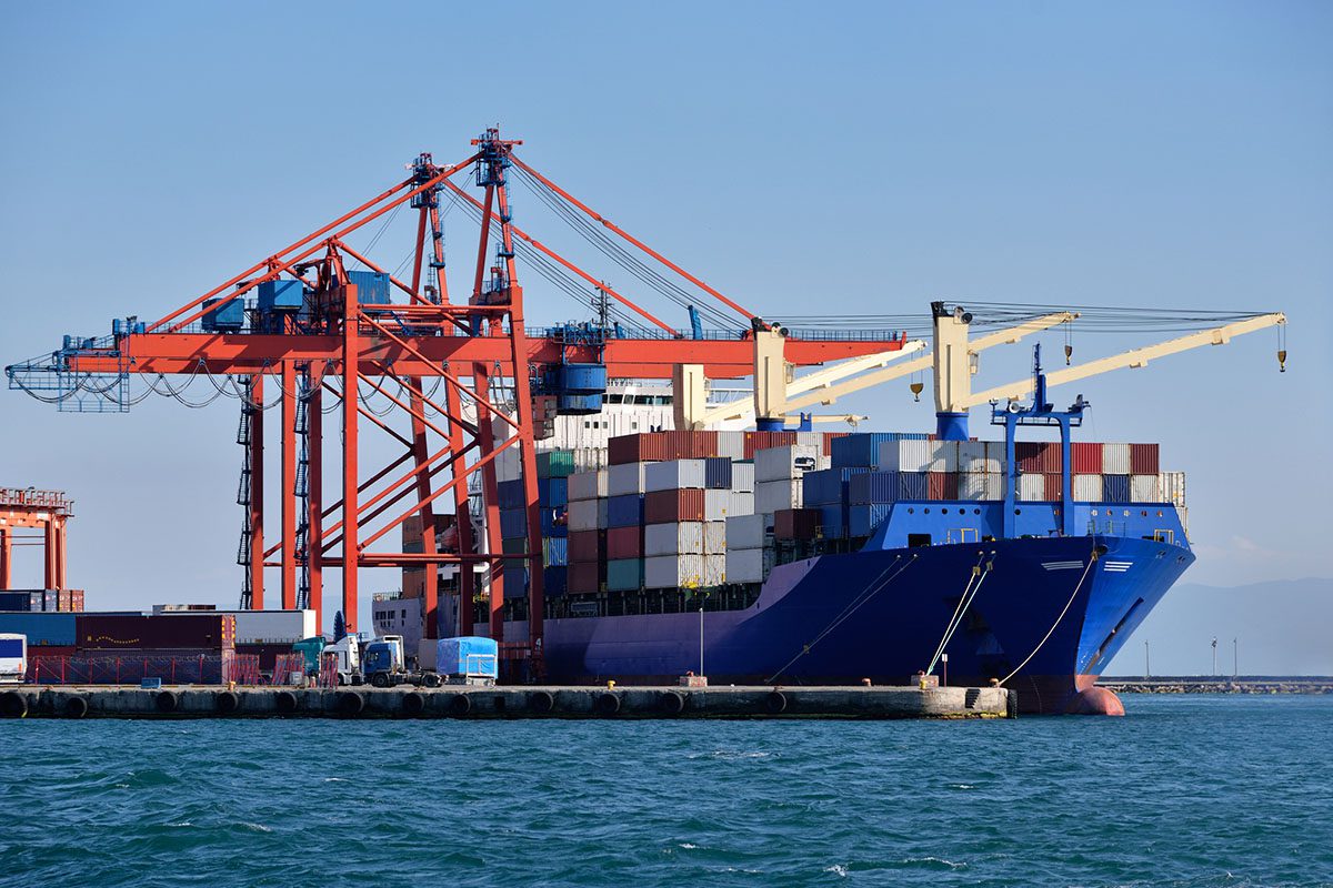 A large cargo ship loaded with shipping containers is docked at a port, alongside red cranes used for loading and unloading containers.