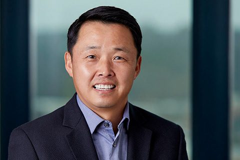 A man in a dark suit jacket and light blue shirt smiles at the camera, standing in front of large windows with a blurred outdoor background.